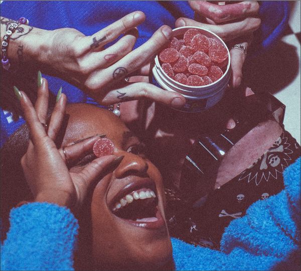 Friends laughing and sharing a tin of red gummy candies; one person holds a gummy over their eye
