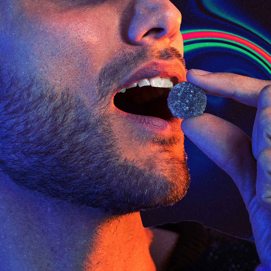 Close-up of a bearded man holding a sugar-coated gummy near his open mouth, blue and orange studio lighting