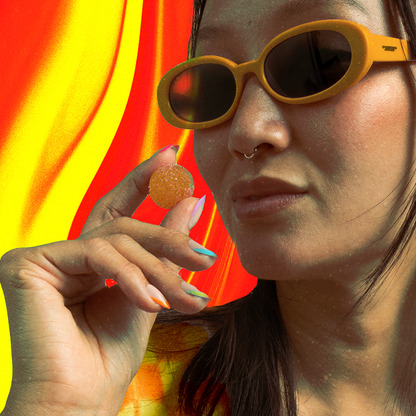 Woman with yellow sunglasses and septum ring holding a sugar-coated gummy between colorful manicured fingers against red-yellow wavy background.